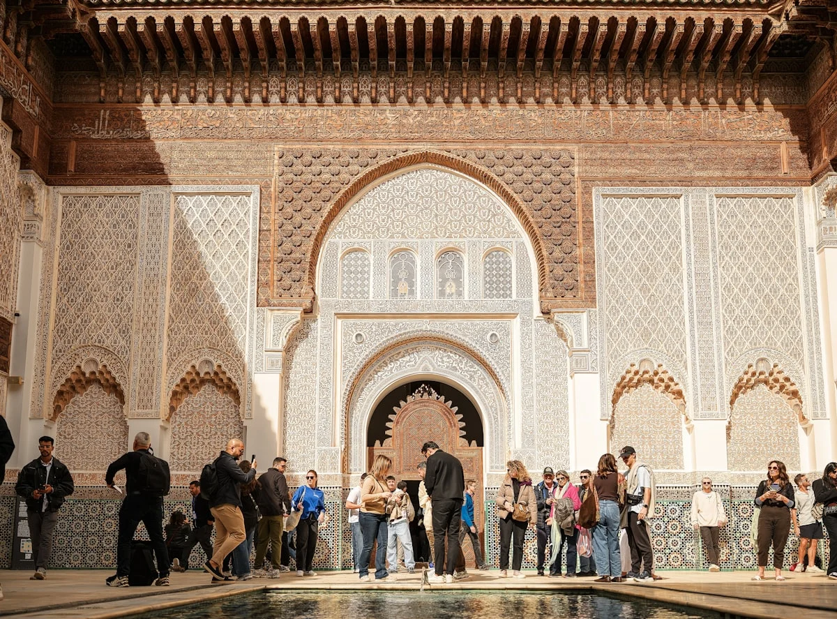 A wide shot of the stunning Moorish architecture inside the Bahia Palace. The frame features meticulously carved stucco walls, cedar wood ceilings, and arched doorways. Tourists wander through the sun-drenched courtyard, providing a sense of scale to the grand historic structures. The warm tones and detailed geometric patterns showcase the peak of Moroccan craftsmanship.