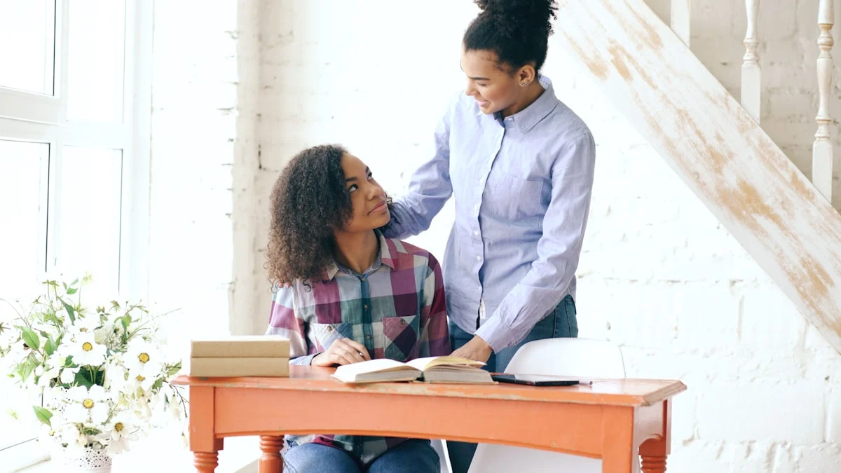 Teenage curly haired mixed race young girl sitting at the table concentrating focused learning lessons and her elder sister helps her studying at home